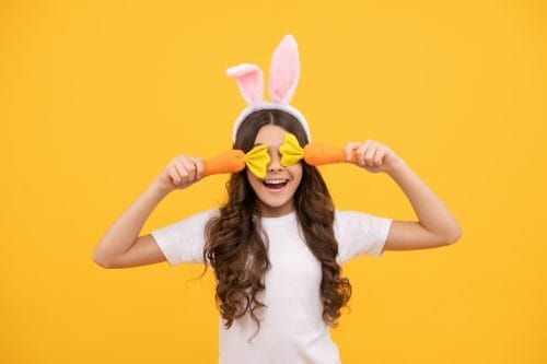 Funny teen girl in bunny ears hold carrot on yellow background, easter.