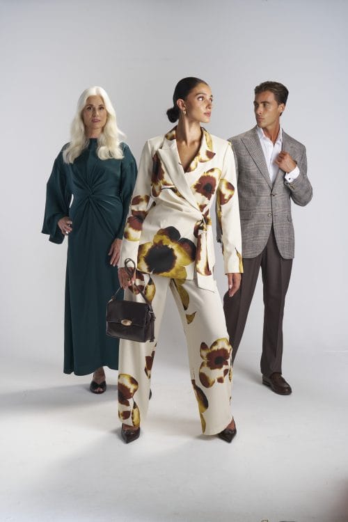 Three models, one woman wearing green overall dress, woman in the middle wearing beige white closed suit and trousers with floral design all over, and man wearing grey and brown closed suit and trousers